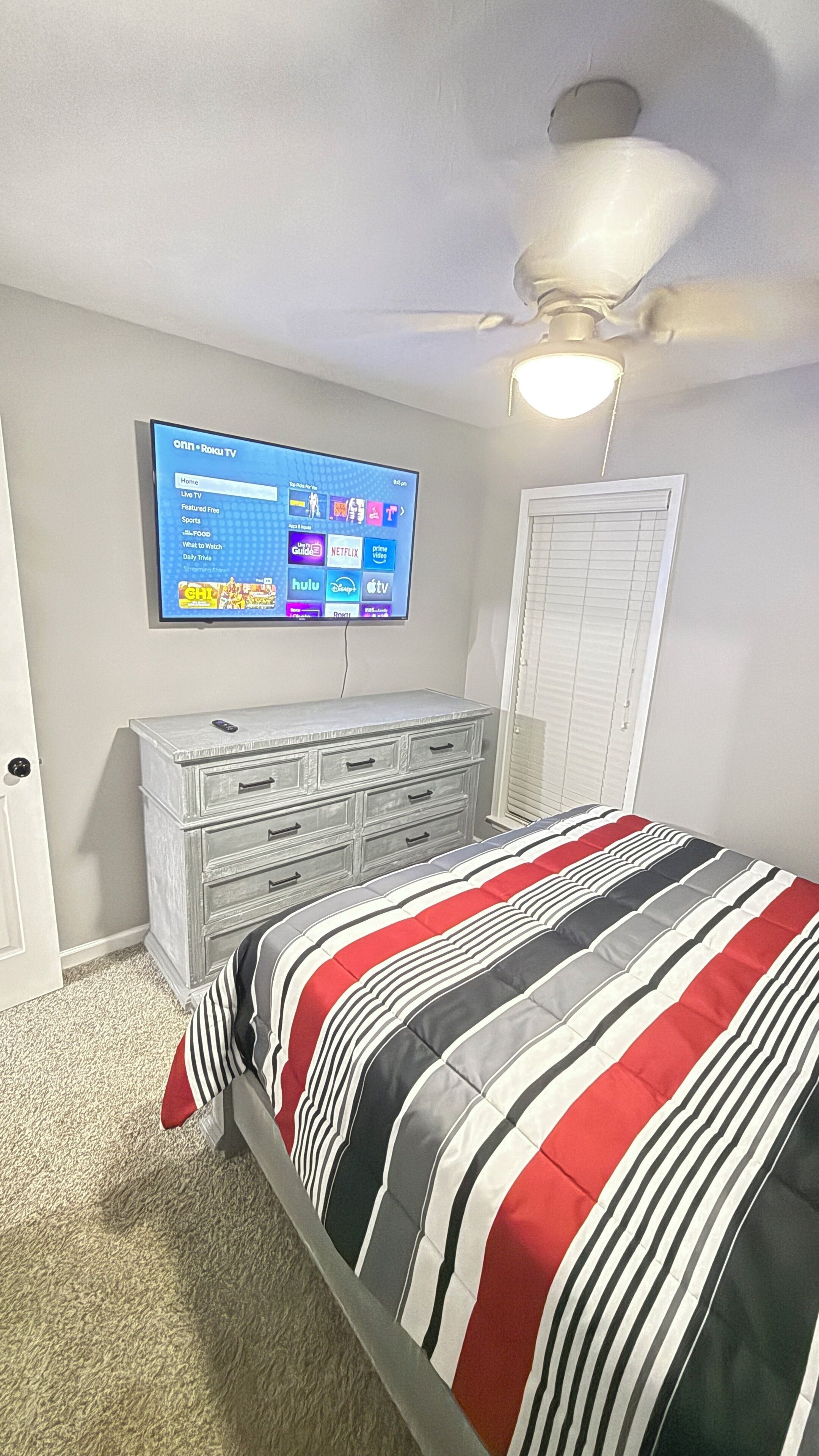 A bedroom with a bed, a dresser, a TV mounted on the wall, and a ceiling fan.