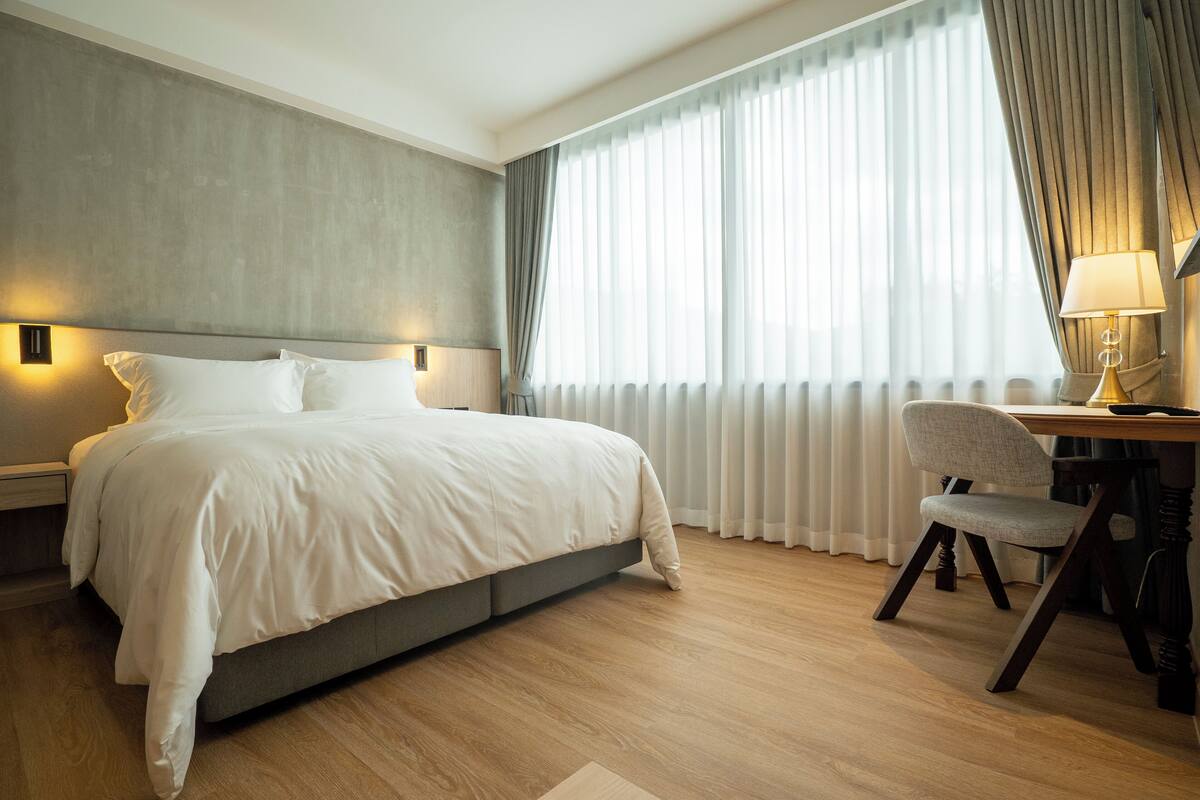 Habitació d'hotel moderna amb un llit gran, una taula de treball amb una cadira i una finestra amb cortines.