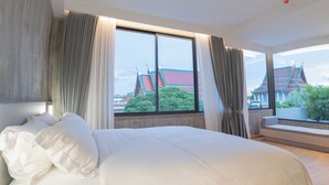 Une chambre d’hôtel avec un grand lit, deux fenêtres avec des rideaux, une vue sur une architecture traditionnelle et un petit balcon agrémenté de plantes.