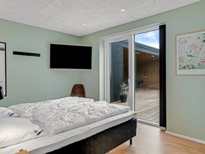 Ett modernt sovrum med en säng, en platt-TV, en stol och en skjutdörr i glas som leder ut till ett utomhusområde.