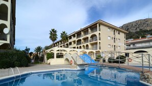 A hotel with a swimming pool, multiple balconies, and a mountain backdrop.