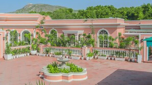 A courtyard with a fountain, surrounded by potted plants and a pink building with arches and columns. - Dolce Vita - Luxury Vacation Villas (Pushkar)