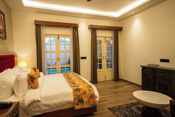 A hotel room with a large bed, a bedside table with a lamp, a wooden headboard, a small round table, and a view of the outdoors through a door.