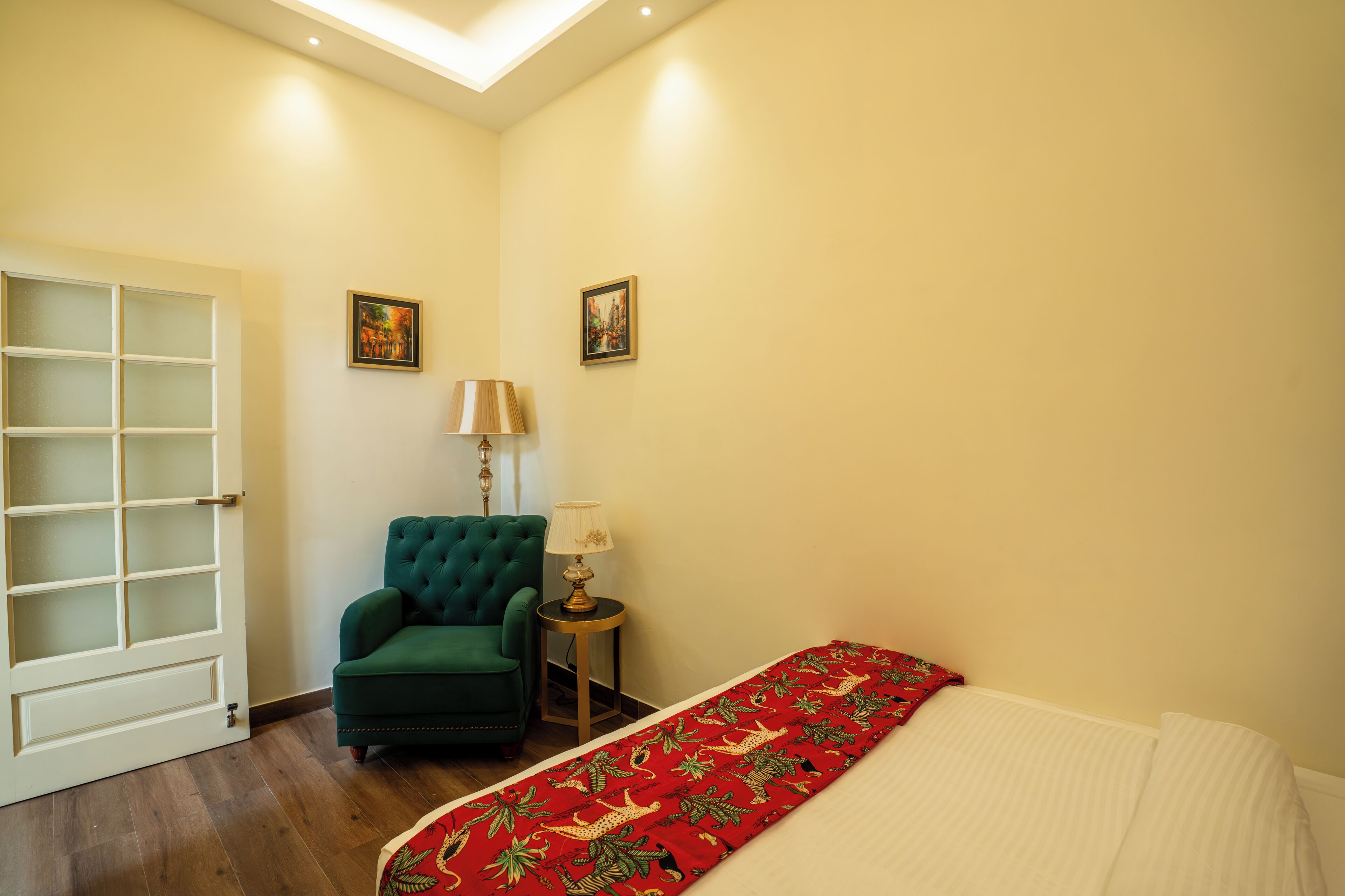 A bedroom with a bed, a green armchair, a small table with a lamp, and two framed pictures on the wall.