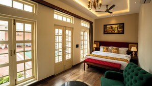 A hotel room with a large bed, a green armchair, a ceiling fan, and a view of the outside through large windows. - Dolce Vita - Luxury Vacation Villas (Pushkar)