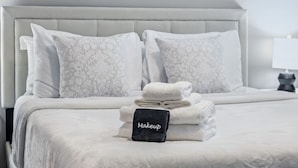 A neatly made bed with a grey headboard, white pillows, and a stack of folded towels with a black tag labeled "Makeup". - Work + Relax | Luxury Escape (Whitestown)