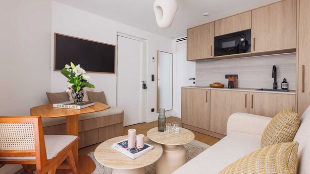 Un salon moderne comprenant un canapé, une table basse, une table à manger avec des chaises, un coin cuisine avec des meubles en bois et un four à micro-ondes, ainsi qu’une grande fenêtre.