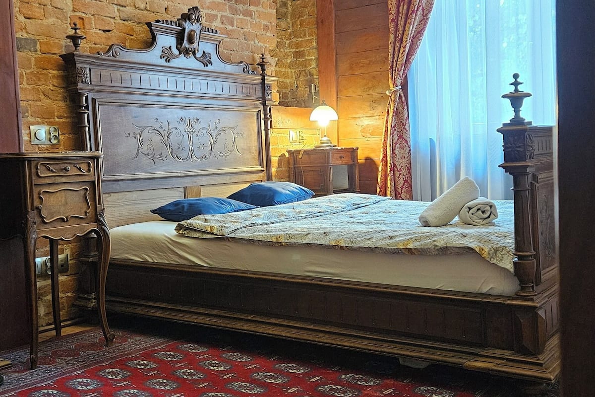Un dormitorio con pared de ladrillo, una cama grande con cabecera de madera, un tapete rojo estampado y una araña.