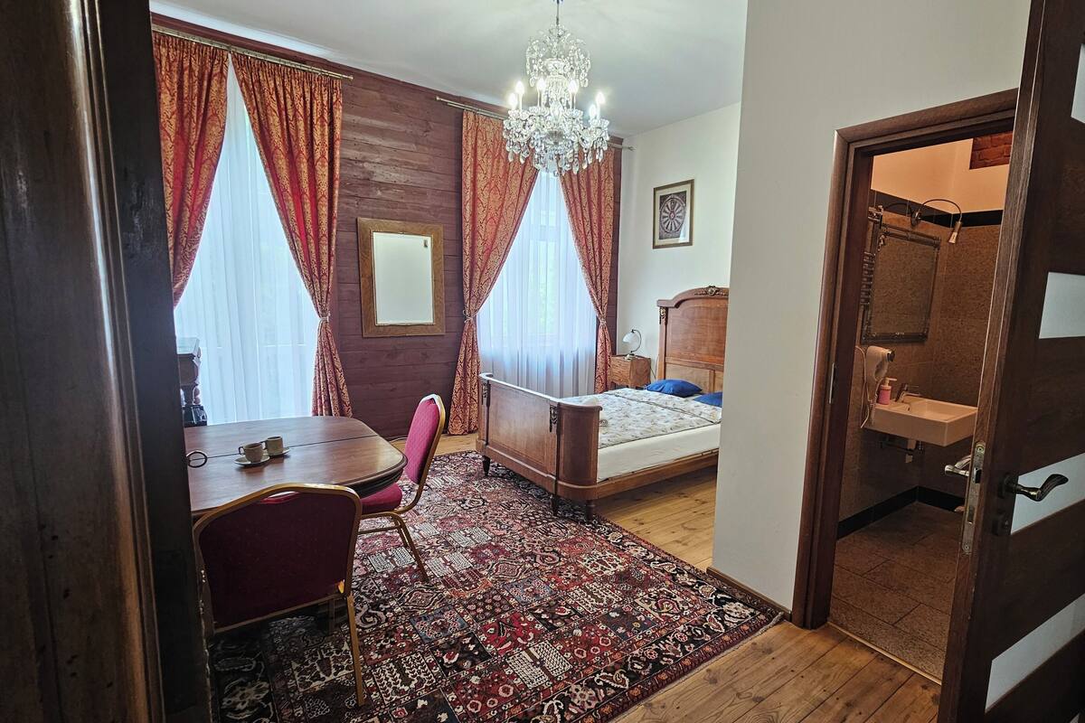Una habitación con una cama, un escritorio y una silla. Hay un candelabro, un espejo y una puerta que da al baño.
