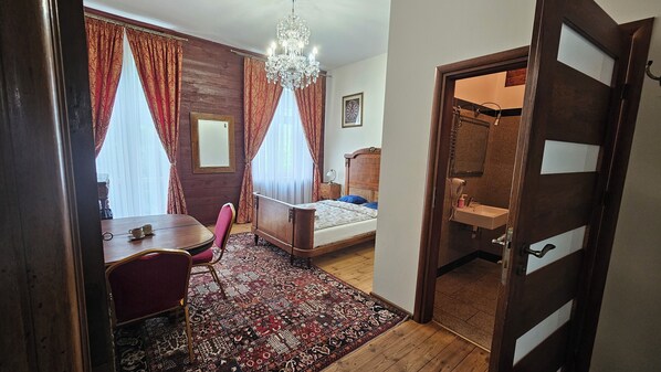 A room with a bed, a desk, and a chair. There is a chandelier, a mirror, and a door leading to a bathroom. - Łódzki Pałacyk Historic Hotel (Lódz)