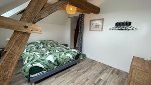 Ein Schlafzimmer mit schrägen Wänden, Holzbalken, einem Bett mit blattgemusterten Überwurf, einem kleinen Nachttisch, einem Wandregal und einem Fenster mit Vorhängen.
