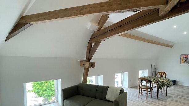 Ein großzügiges Loft mit hohen Decken, freiliegenden Holzbalken, einem grünen Sofa, einem Esstisch mit Stühlen und einem gemusterten Teppich.