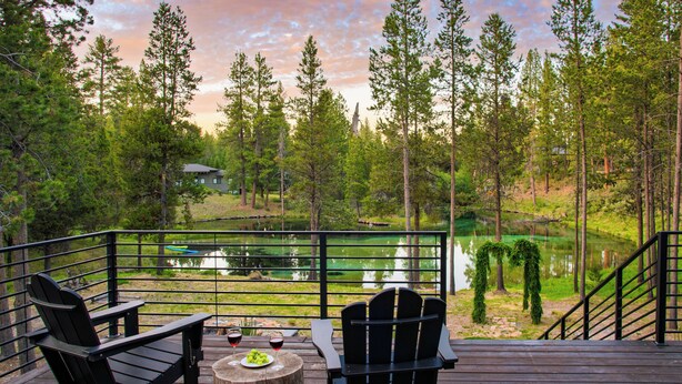 Una terraza con dos sillas Adirondack, una mesa redonda de madera y vista a un lago rodeado de árboles.