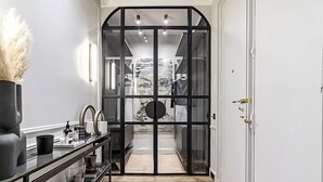 Un pasillo con una puerta de vidrio de marco negro que da al baño. Hay un tocador con espejo, un jarrón con plantas secas y un estante con diversos objetos.