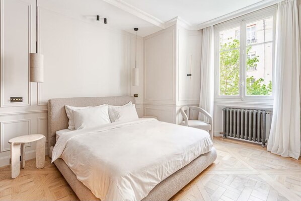 A bedroom with a large bed, a chair, a small stool, and a radiator.