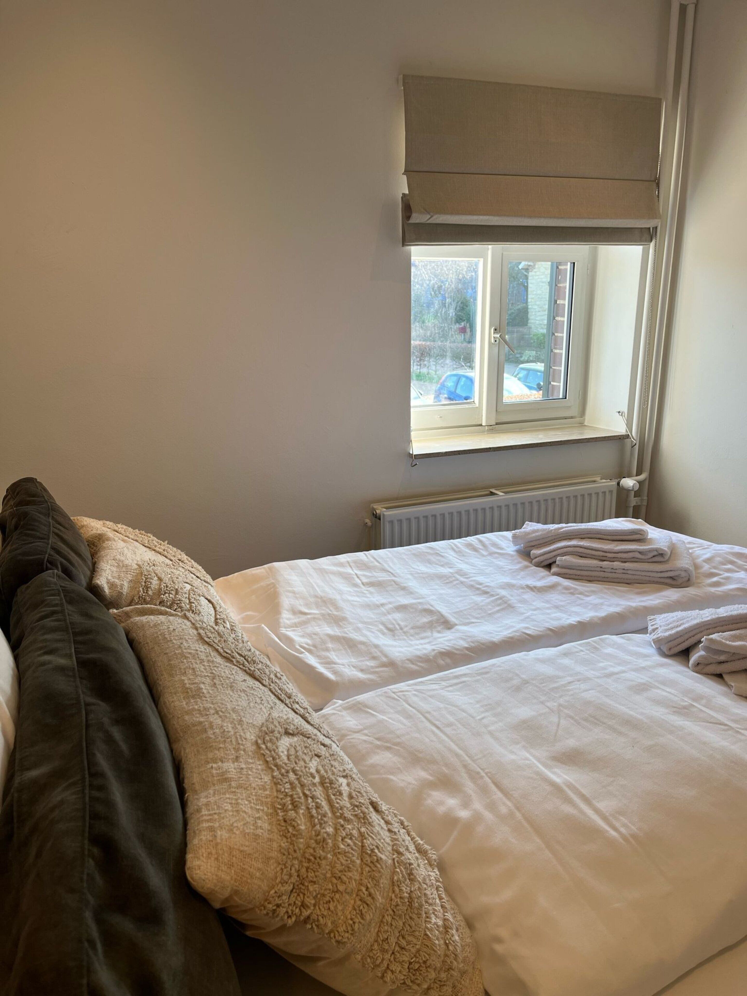 Egy hálószoba két ágyjal, radiátorral, egy ablakkal, melyből kilátás nyílik a külvilágra, és egy mennyezeti ventilátorral.