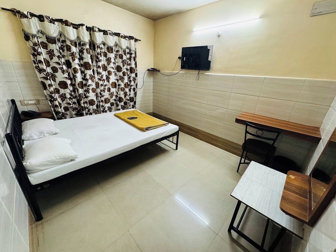 A small, clean room with a single bed, a desk, and a chair. There is a curtain with a floral pattern, a wall-mounted TV, and a power outlet.