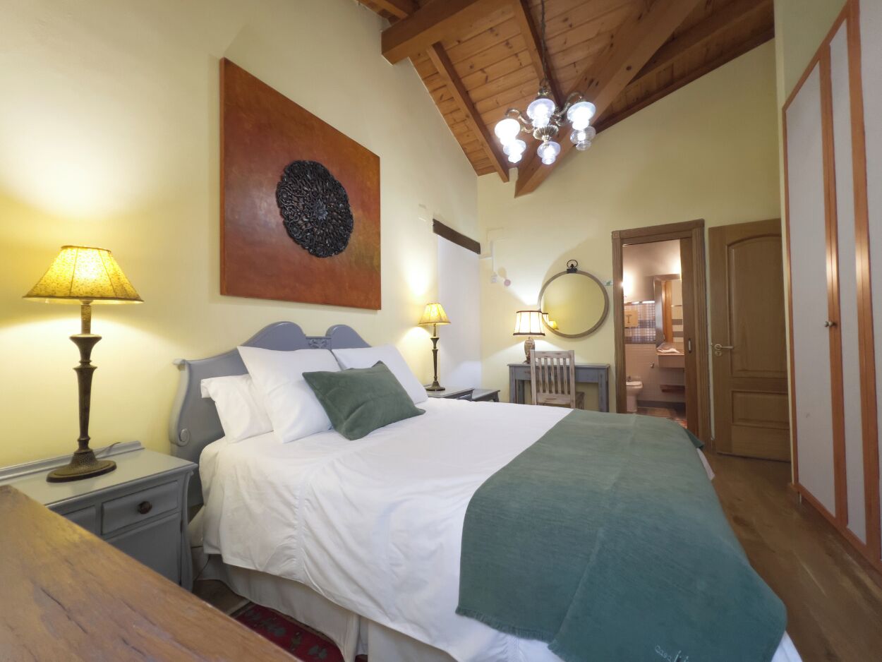 Un dormitorio con cama, dos mesitas de noche con lámparas, techo de madera, espejo en la pared y puerta que da al baño.