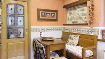 Une cuisine chaleureuse avec une table en bois, des chaises et un banc agrémenté d’un coussin à motifs floraux. Un élément décoratif est accroché au mur, au-dessus du banc.