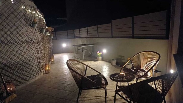 Ein Terrassenbereich im Freien mit Korbstühlen und einem kleinen runden Tisch, beleuchtet durch Laternen und Lichterketten.