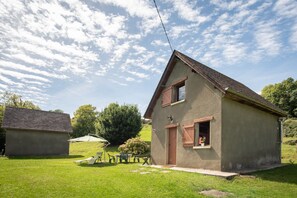 Una casa pequeña con techo inclinado, una puerta principal y una ventana con persianas.  Hay un jardín con una mesa y sillas, y un árbol al fondo.