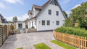 A two-story house with a steeply pitched roof, a stone foundation, and a paved driveway leading to a wooden gate. - Carlotta, Germany (Sylt-Ost)