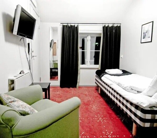 Une chambre d’hôtel avec un lit, un fauteuil vert, une télévision et une moquette rouge.