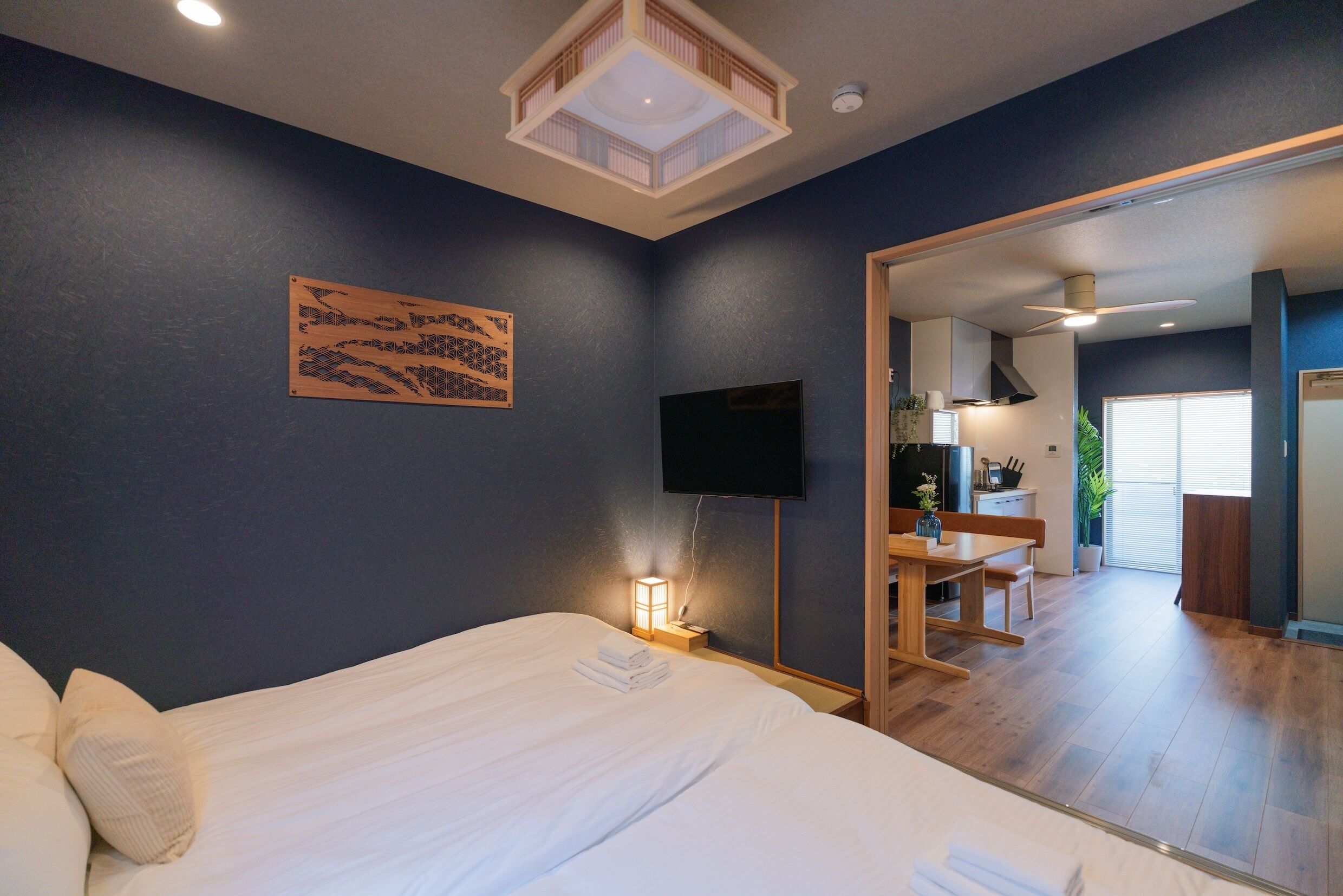 A hotel room with a large bed, a flat-screen TV, a bedside table with a lamp, a wooden artwork on the wall, and a view into the kitchen area.