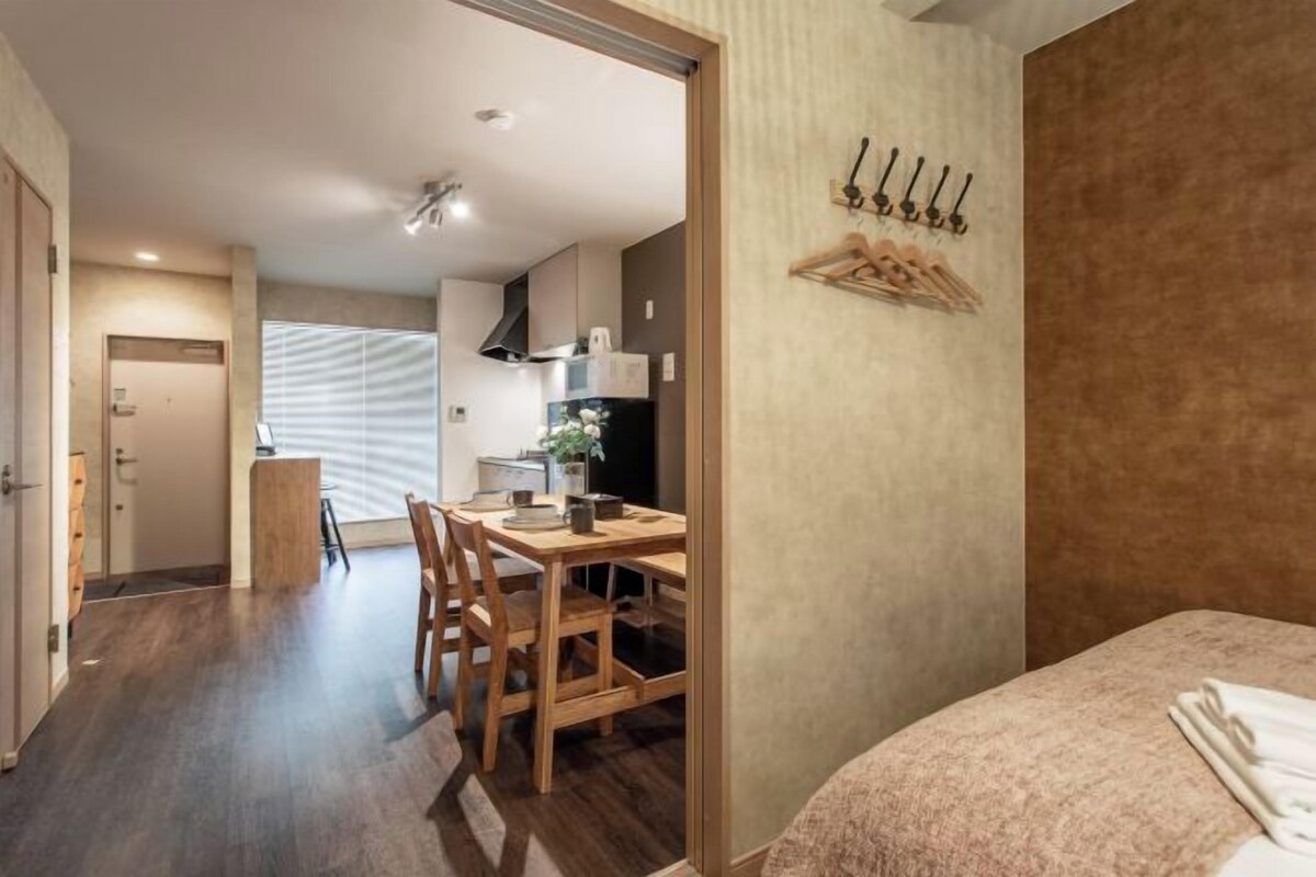 A hotel room with a bed, a wooden dining table with chairs, a kitchen area with a microwave, and a coat rack.