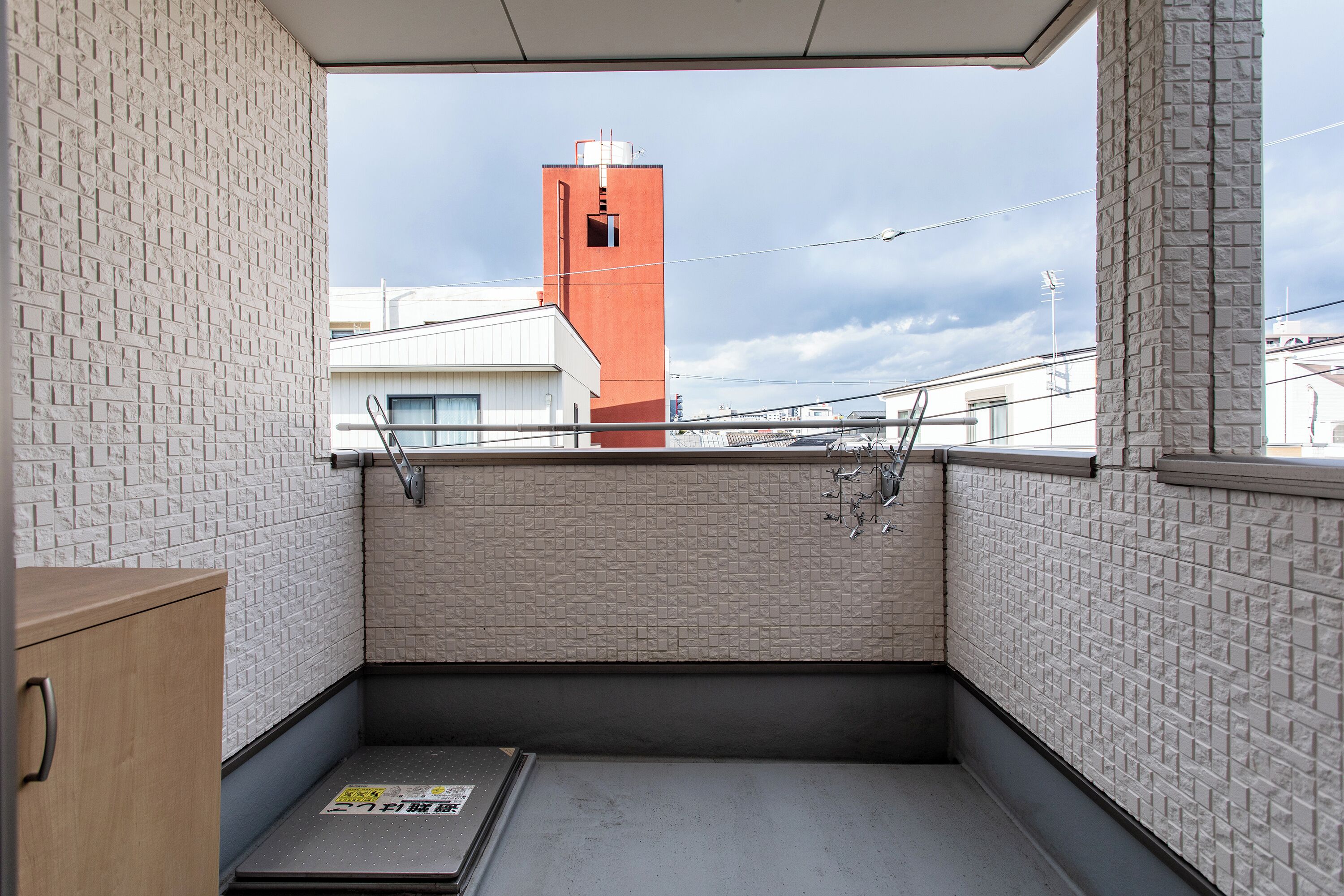 A small balcony with a view of a red tower, white brick walls, and a metal ladder.