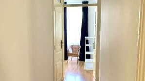 A narrow hallway with wooden flooring, white walls, and a door leading to a room with a wicker chair and a white shelf. - River view over Tage (Lisboa)