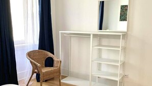 A bedroom with a white bed, a wicker chair, a white desk with shelves, and a window with curtains. - River view over Tage (Lisboa)