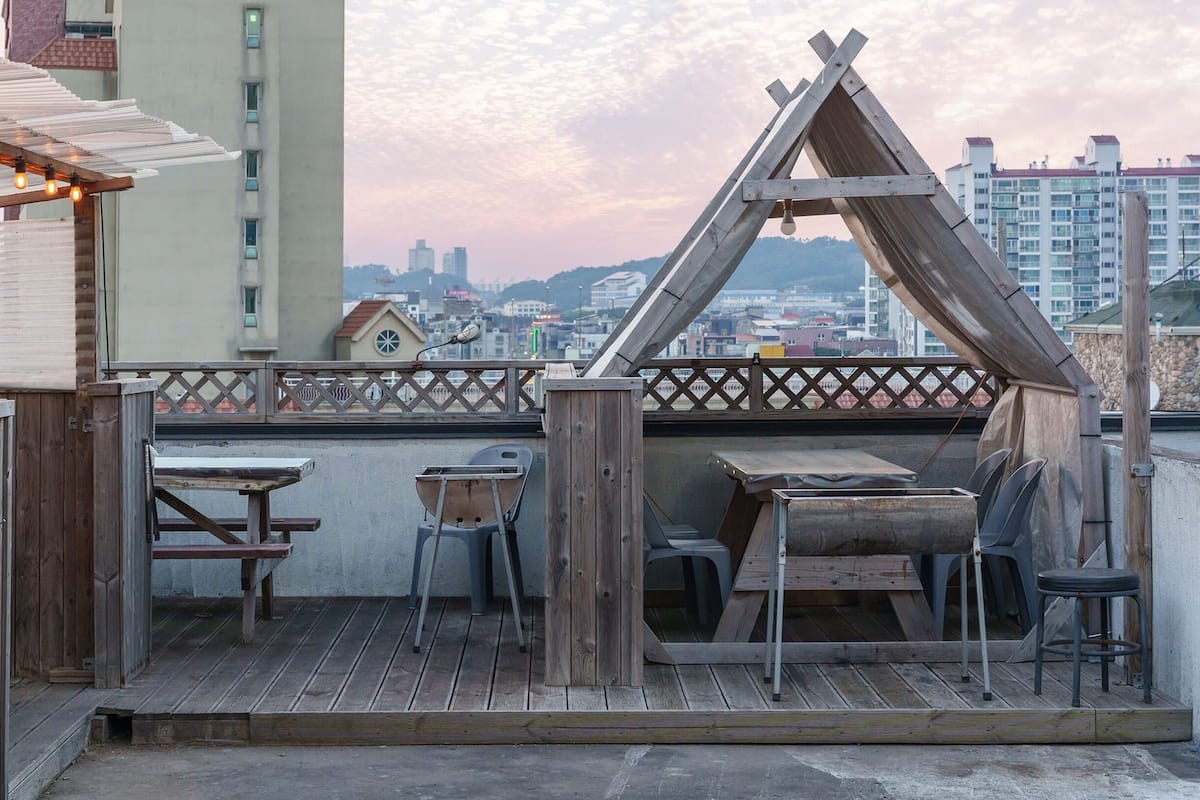 A rooftop terrace with wooden furniture, a wooden structure resembling a teepee, and a view of the cityscape.