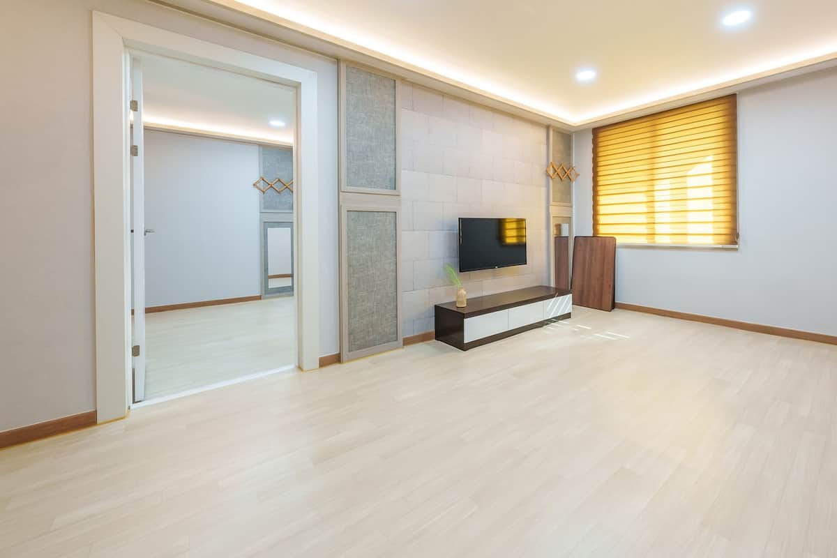 A modern living room with a flat-screen TV mounted on a textured wall, a wooden entertainment unit, and a large window with blinds.