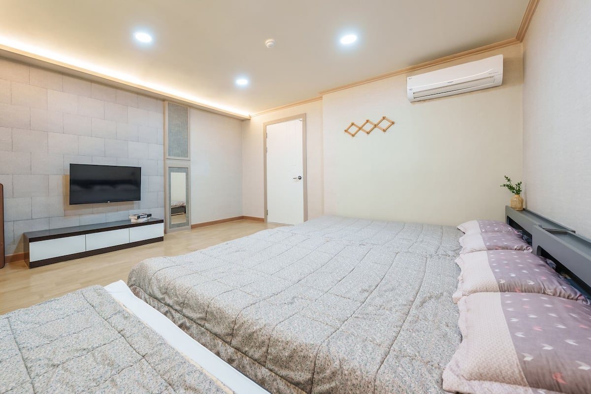 A modern bedroom with a large bed, a TV mounted on the wall, and a built-in air conditioning unit.
