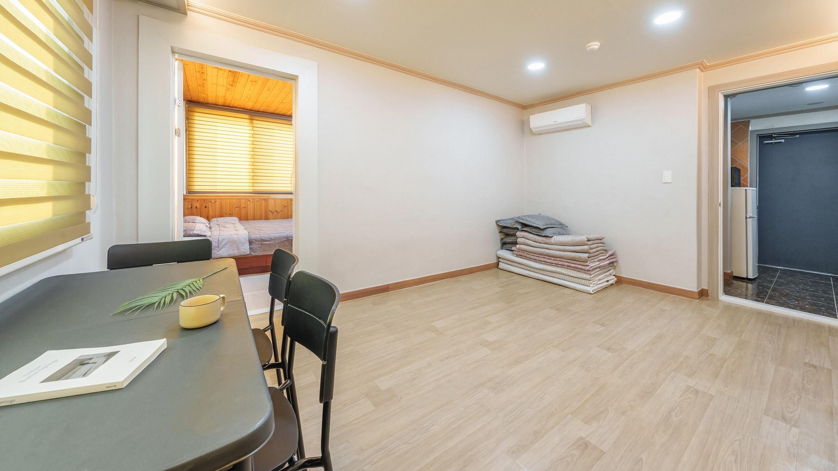 Une pièce comprenant une table à manger, des chaises, un lit avec une tête de lit en bois, une fenêtre avec des stores, une pile de serviettes pliées et un appareil de climatisation.