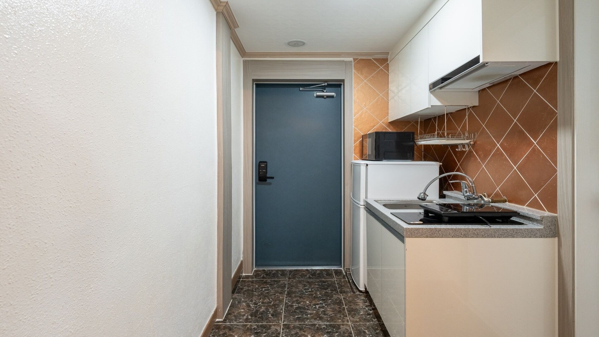 A narrow hallway leading to a door with a key lock, a small kitchen area with a sink and a microwave, and a tiled backsplash.