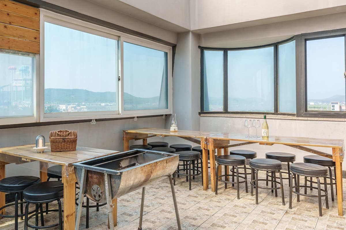 A rooftop bar with multiple tables and stools, large windows offering city views, and a grill.