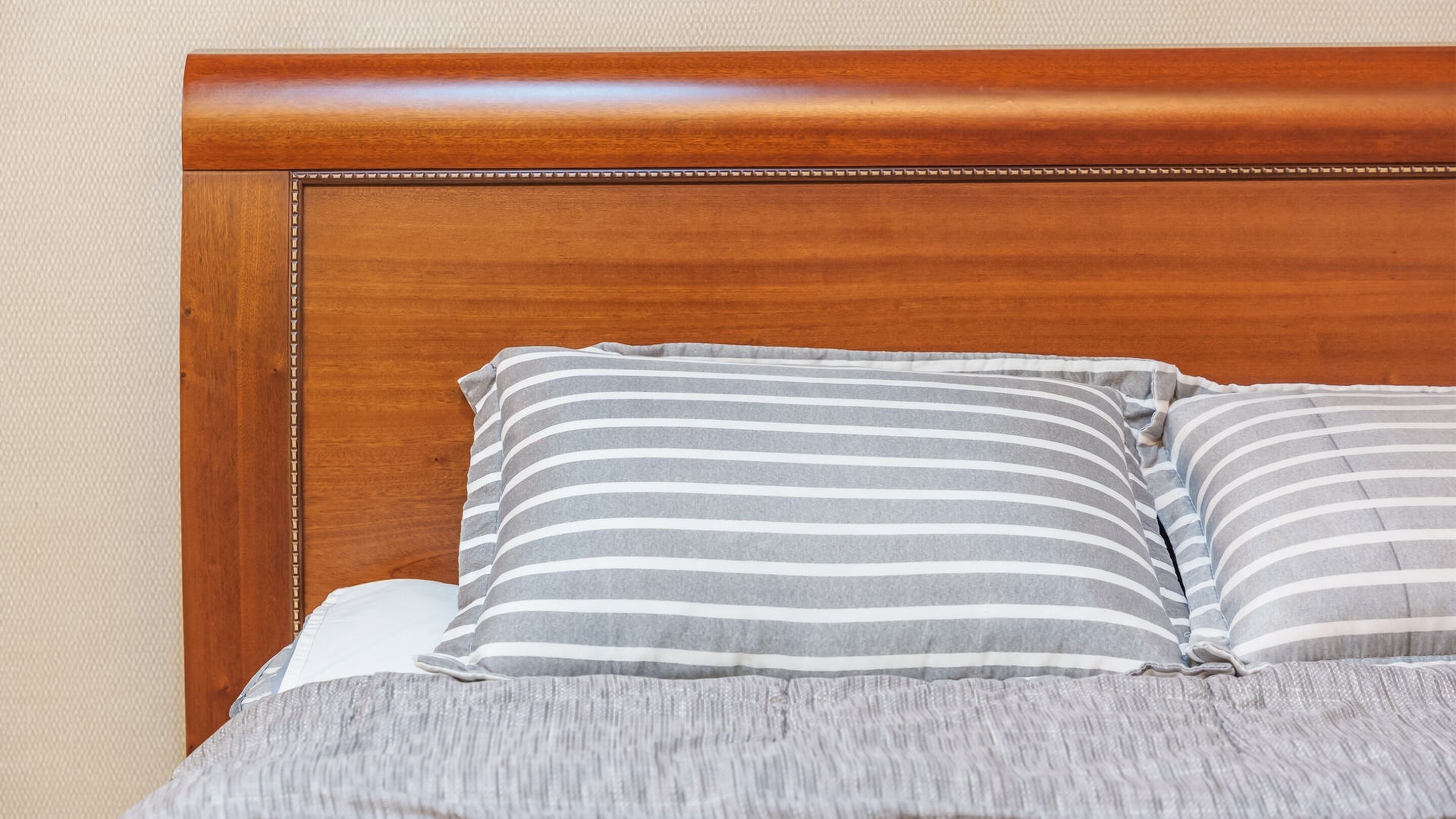 Una cama con cabecera de madera, dos almohadas a rayas y una manta gris.