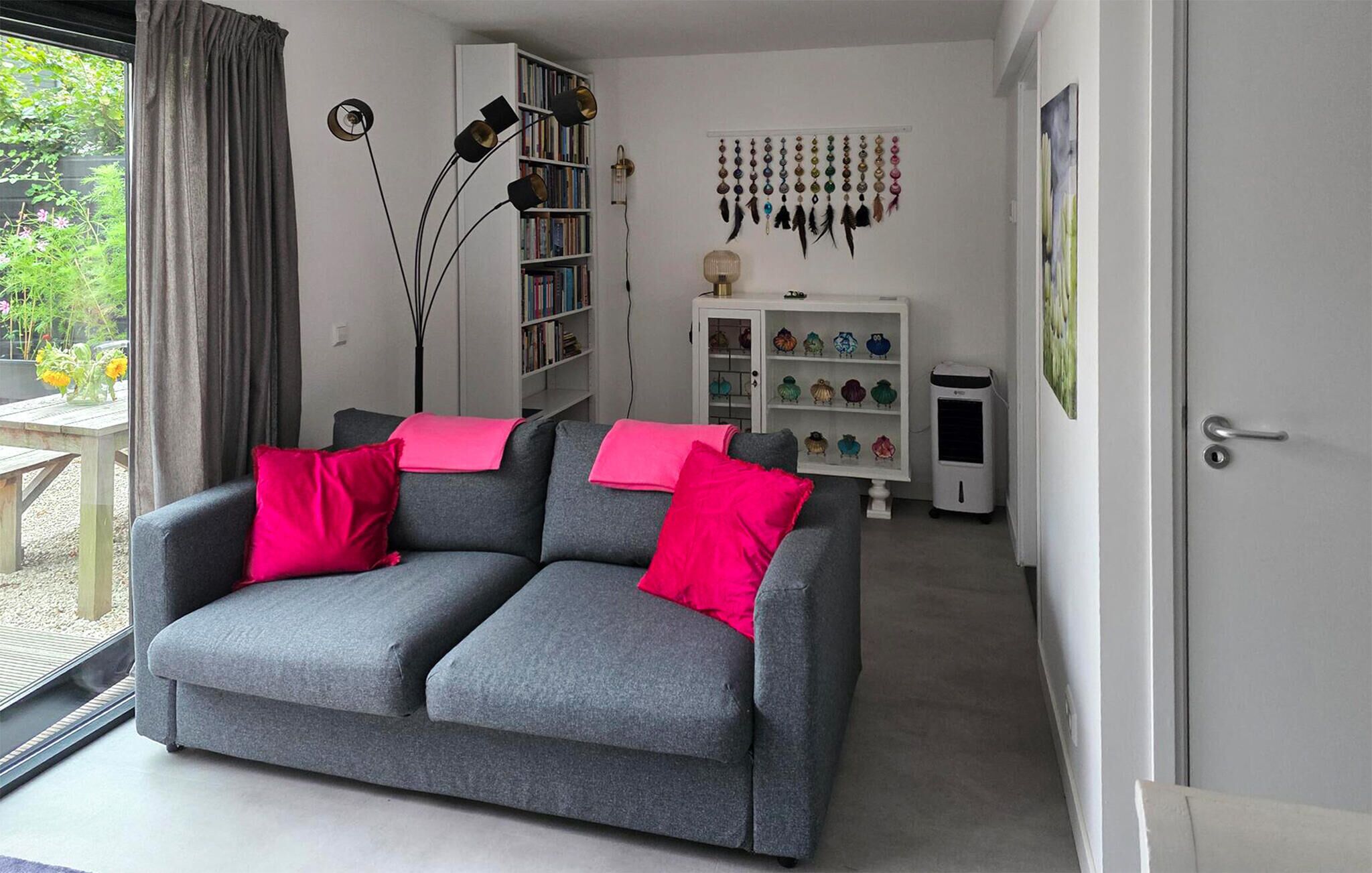 Ein modernes Wohnzimmer mit einem grauen Sofa, roten Kissen, einem Bücherregal, einem Wandbehang und einer weißen Kommode mit bunten Gegenständen.