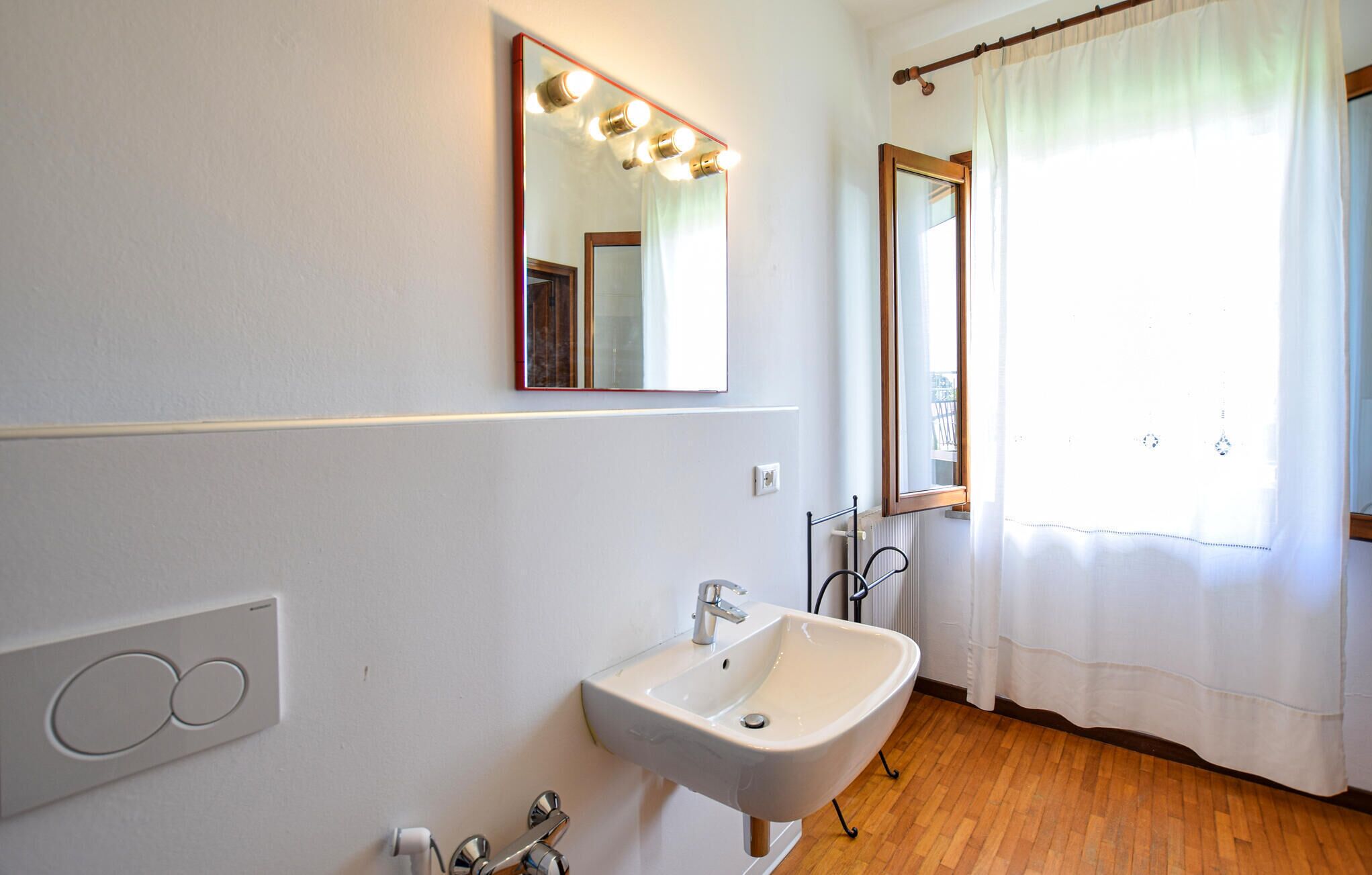 A bathroom with a white sink, a mirror with lights, a window with a curtain, and a radiator.