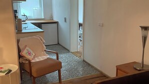 Ein kompakter Wohnraum mit Küchenzeile, einem Sessel mit Kissen, einem kleinen Beistelltisch und einem Bett.