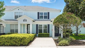 A two-story residential building with a light blue exterior, dark shutters, and a well-maintained front yard with hedges and a tree. - Disney Escape - Private Pool & Full Resort Access (Kissimmee)