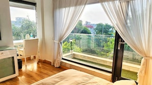 Une chambre avec un grand lit, un bureau et une chaise, ainsi qu’un balcon offrant une vue sur la verdure et les bâtiments.