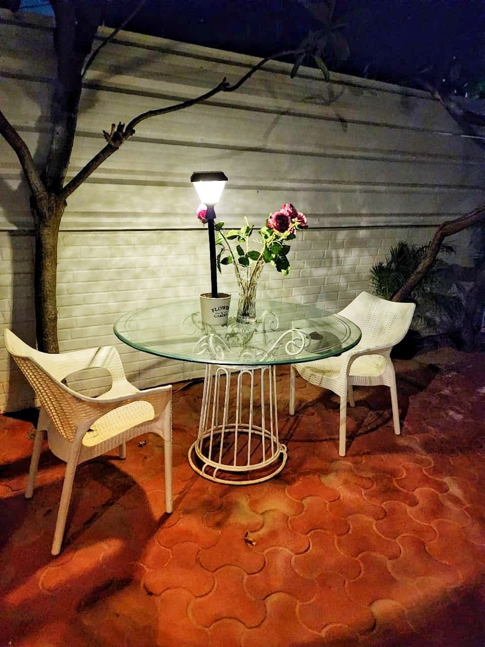 A round glass table with a metal base, surrounded by two white chairs. A small lamp with a flower in a vase is placed on the table.