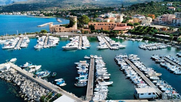 A marina with numerous boats docked, a prominent building with a tower, and a coastal town in the background.