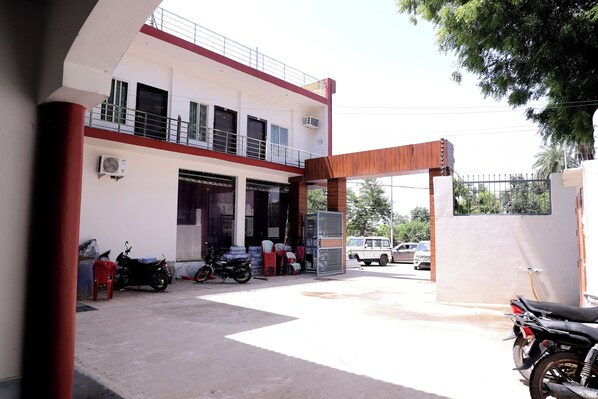 A building with a two-story structure, a covered entrance, and a parking area with motorcycles and a car. - Hotel Murliwala Gopala (Govardhan)
