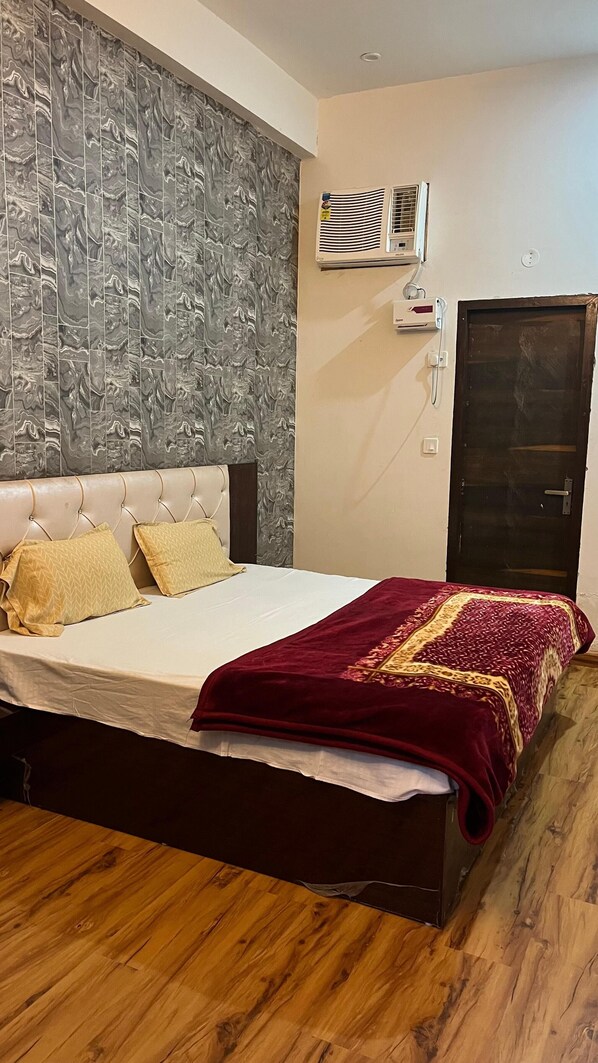 A bedroom with a marble-patterned headboard, a bed with a white bedspread and two pillows, a red throw blanket, and a wooden floor. - Hotel Murliwala Gopala (Govardhan)