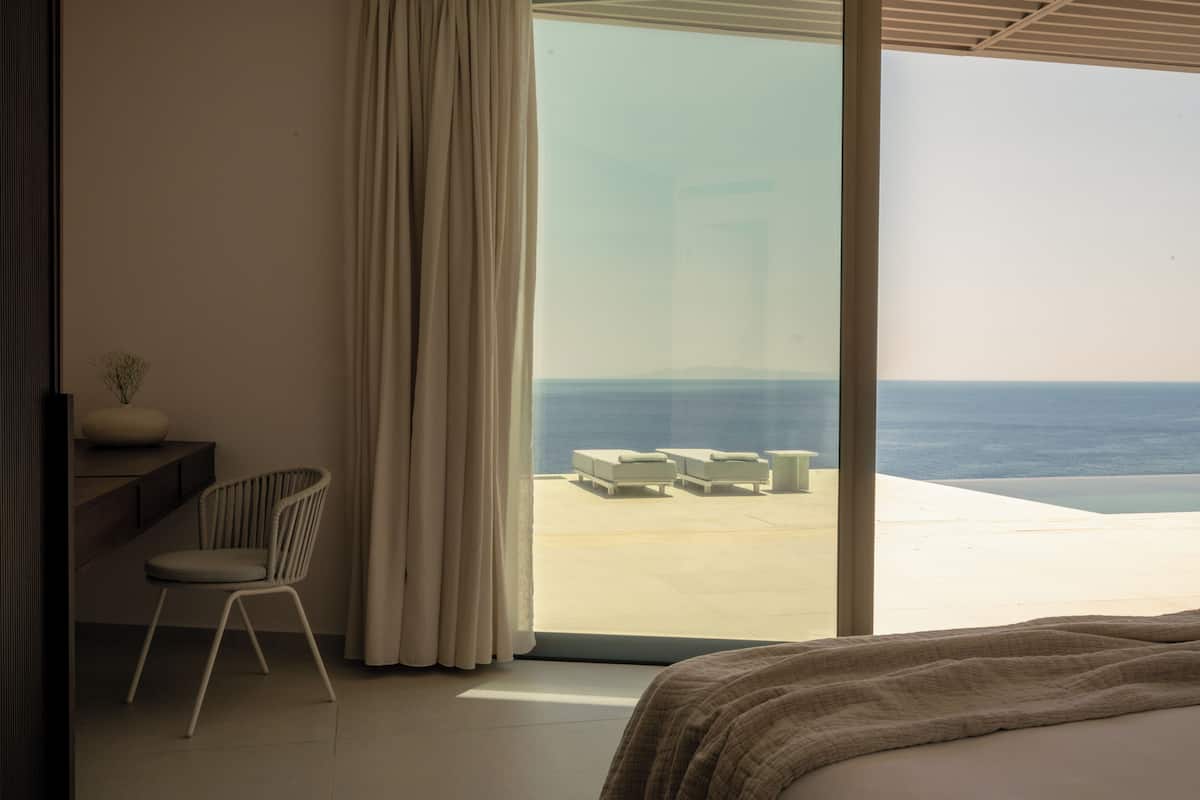 A modern hotel room with a large window, a bed, a chair, and a small table.