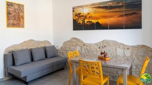 Una habitación con pared de piedra, un sofá gris, una mesa con sillas amarillas y dos obras de arte en la pared.